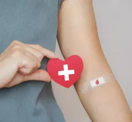 A close up of a person's elbow. Their arm is covered with a bandage with an image of a drop of blood on it. The person is holding a heart with a white cross in the center.