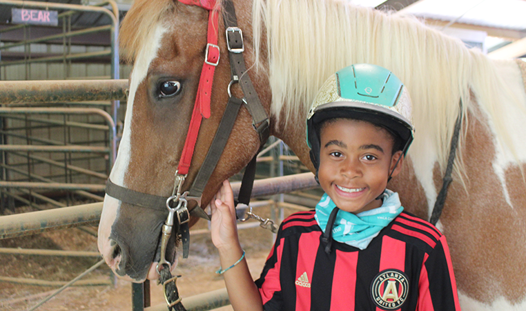 A boy wearing a riding helmet smiles at the camera. He is standing beside a horse.