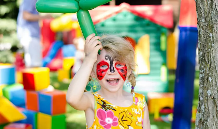 Child with face painting holding a balloon animal in front of bounce house. 