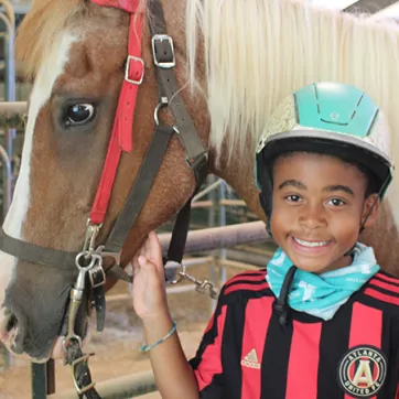 A boy wearing a riding helmet smiles at the camera. He is standing beside a horse.