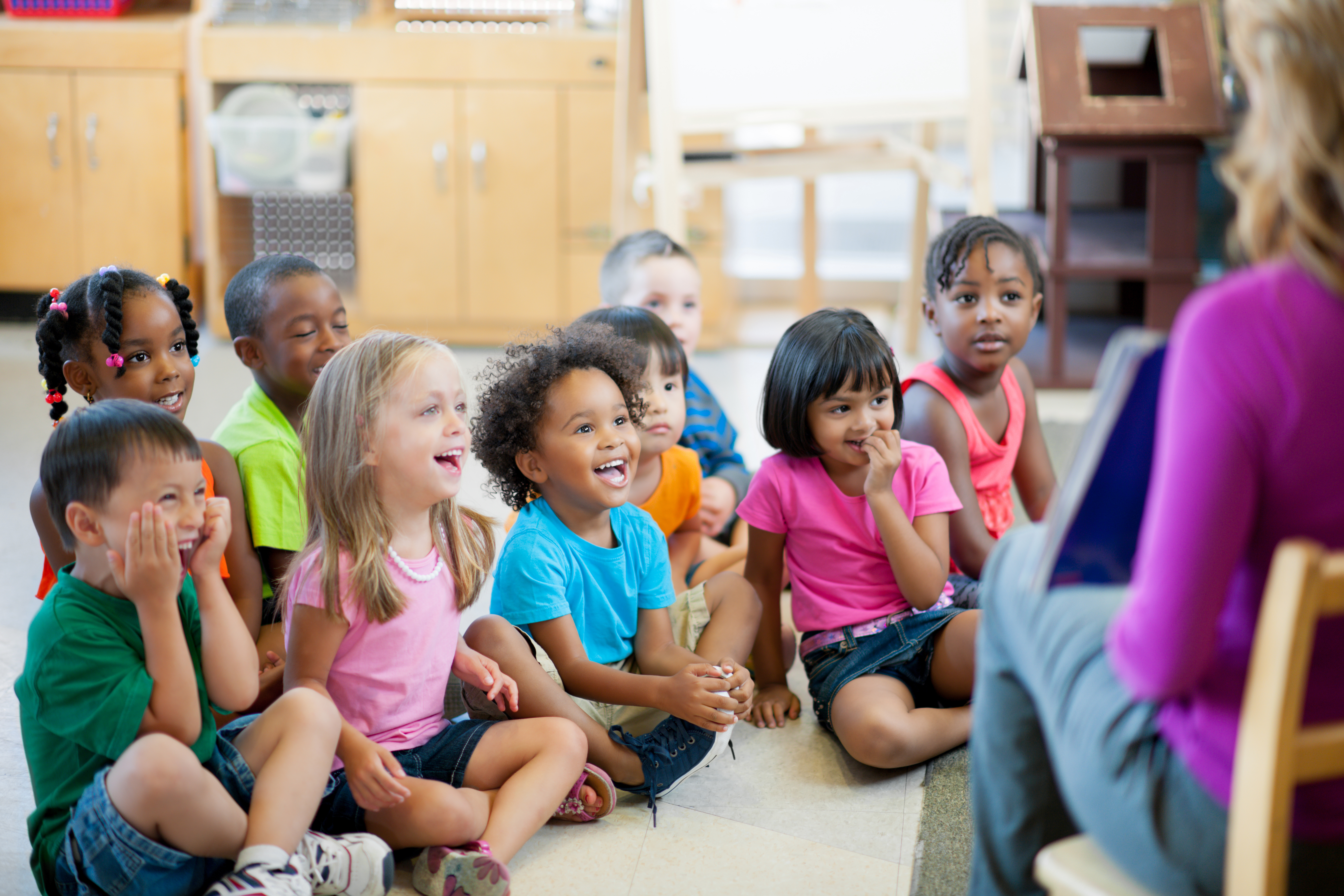 A diverse group of children sitting together listening to a teacher read.
