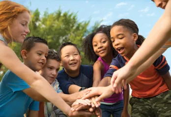 A group of diverse children at summer camp stand in a circle. Their hands are stacked in the middle of the circle.