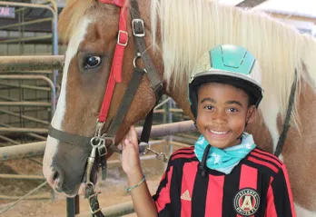 A boy wearing a riding helmet smiles at the camera. He is standing beside a horse.