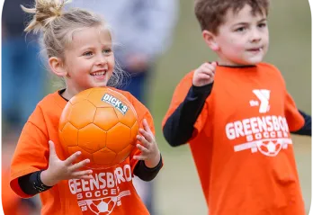A young boy and girl wearing orange shirts that say Greensboro Soccer in white text. The girl is holding an orange soccer ball and smiling.