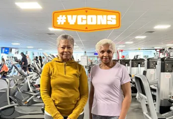 Two women standing in a ymca wellness center, smiling for a photo. Graphic #YCON above their head.