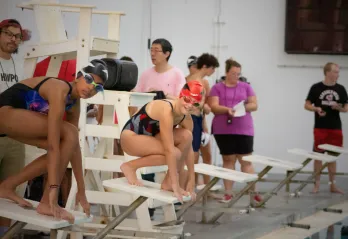 Two swimmers wearing swim caps and goggles prepare to race from starting blocks at an indoor pool during a GCY Makos swim meet.