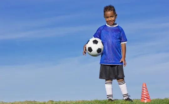 A young boy smiles at the camera while holding a soccer ball at his side.