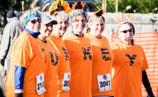 A group of women standing together. They are each wearing orange shirts with a letter on them. The shirts spell out "tirkey."
