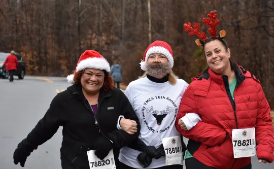 A group of women smiling at the camera. One woman is wearing a Santa hat, while another wears reindeer antlers. Another woman is wearing a Santa hat and beard.