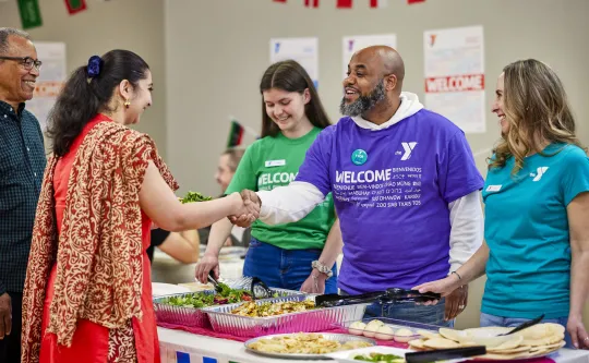 Three Y team members greet a visitor to the Y. She is shaking hands with a man in a purple YMCA shirt with the word 'Welcome' in different languages.