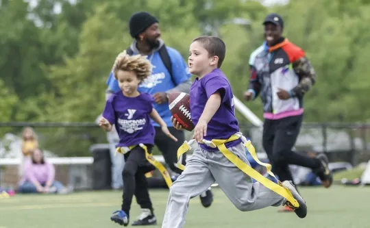 Children playing flag football at the Bryan YMCA. Two Y team members are running alongside a young boy carrying the ball.