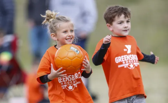 A young boy and girl playing soccer at the Spears YMCA. They are wearing bright orange shirts. The girl is holding an orange soccer ball.