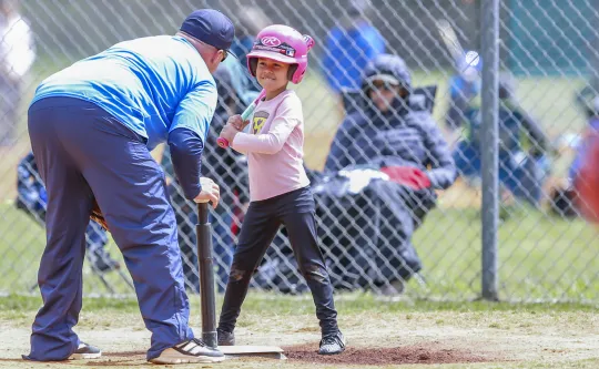 A coach leans down to speak with a young girl at a t-ball/baseball game. The girl is smiling and wearing a pink helmet.