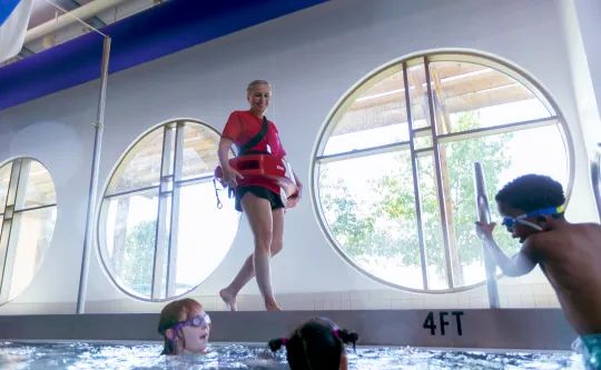 A lifeguard walking along the side of a pool. She is smiling at a group of children swimming.