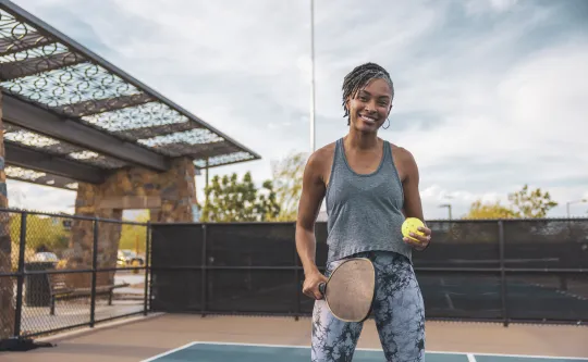 A woman holding a pickleball and pickleball racquet standing in a court outdoors. She is smiling at the camera.