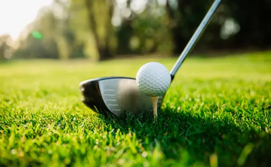 A close-up image of a golf ball on a tee. A golf club is poised near it.