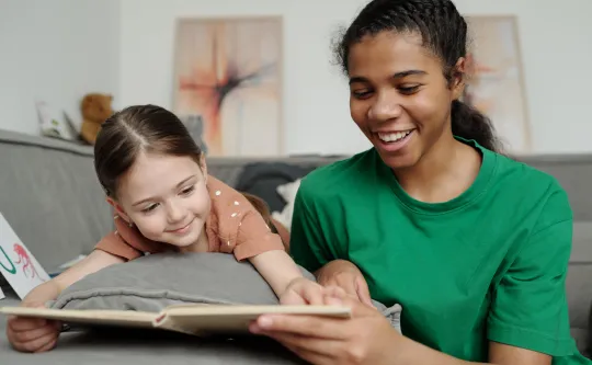 A teenage girl reads to a young girl. They are both smiling.