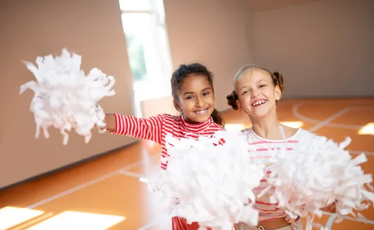 Two young cheerleaders standing together in the gym. They are both holding white cheerleading pom-poms.