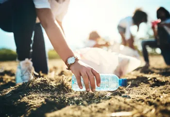 Someone in the foreground is leaning down to pick up a plastic bottle from the ground. In the background, several other people are cleaning up litter.