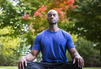 A man sitting in the grass outside on a yoga mat. He has his eyes closed and is sitting cross-legged in a yoga pose.