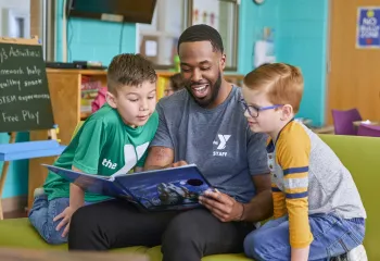 A Y team member reads with two young boys during the YMCA's afterschool program. He is smiling while they look at the open book together.