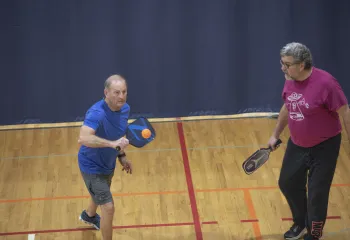A pickleball game at the Ragsdale YMCA. Two men on a team stand together. The man on the left is sending a bright orange ball back to their opponent.