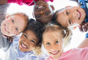 A diverse group of children laughing and playing. They are standing in a circle together and looking down at the camera.