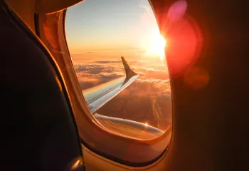 An interior view of an airplane. The plane's wing and a sunset over the clouds can be seen through a window.