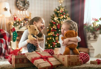 Two sisters open gifts together on Christmas morning. They are both holding stuffed bears.