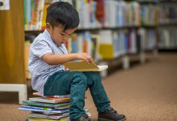 A young boy reading a book. He is sitting on a stack of children's books.