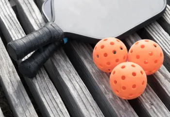 Two pickleball paddles and three pickleballs sitting on a wooden surface.