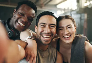 A group of three young adult friends at the gym. They're smiling into the camera.