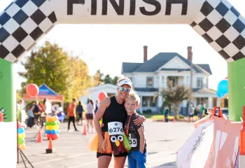 A woman wearing sunglasses and a turkey-themed costume and a young boy stand together outside. They are standing under the road race arch together and smiling at the camera.