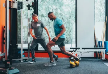 A personal trainer instructs his client in exercise forms and techniques in a gym with orange walls, gray carpet, and large windows. The men are both wearing athletic clothing.