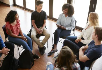 A diverse group of teens sitting together in chairs arranged in a circle. The group is listening to a young woman speak.