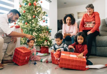 A multigenerational family sitting together on Christmas morning. They are watching three young children open presents.