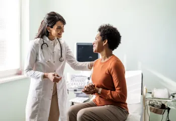 A doctor speaking to a patient in a medical setting. The doctor and patient are both smiling, and the doctor has her hand on her female patient's shoulder.