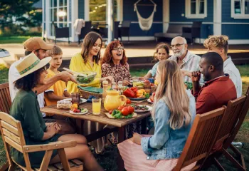 A diverse group of people eating outdoors. They are sitting at a wooden table in wooden folding chairs. A grill is nearby to their left.