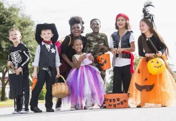 A diverse group of children wearing Halloween costumes. They are standing outside in a group and holding trick or treating bags and buckets.