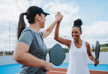 Two women holding Pickleball racquets outdoors. They are smiling and high-fiving each other.
