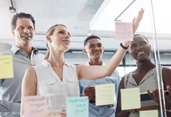 A diverse group working on a vision board together in the form of Post-Its on a glass wall.