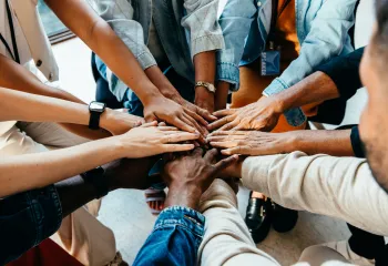 A diverse group of people standing in a circle. They are placing their hands in the center of the group.