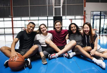A diverse group of teenagers sitting together on a padded floor mat in a gym. On the left side of the group, a young teen has his hand on a basketball in front of him.