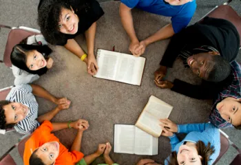Overhead view of a diverse group of people sitting around the table, with open bibles in the center looking up at the camera smiling.