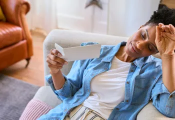 A woman sits on a couch, fanning herself with a card and looking flushed, illustrating a hot flash.