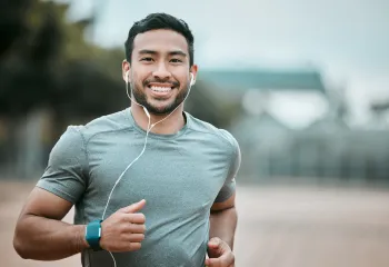 A man running outdoors while wearing earbuds. He is smiling at the camera.