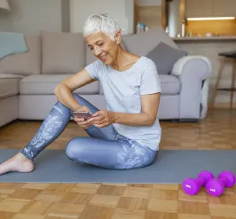 A senior woman works out at home. She is sitting on a yoga mat while using her phone.