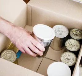 A photo of canned goods inside a cardboard box. There is a man's arm reaching to pick up a can.