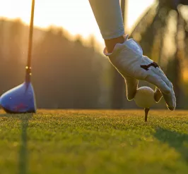 A gloved hand adjusts a golf tee on the golf course. There is a bright sunset and a golf club in the background.