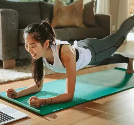 A woman working out at home smiles at her laptop. She is on a yoga mat in her living room.